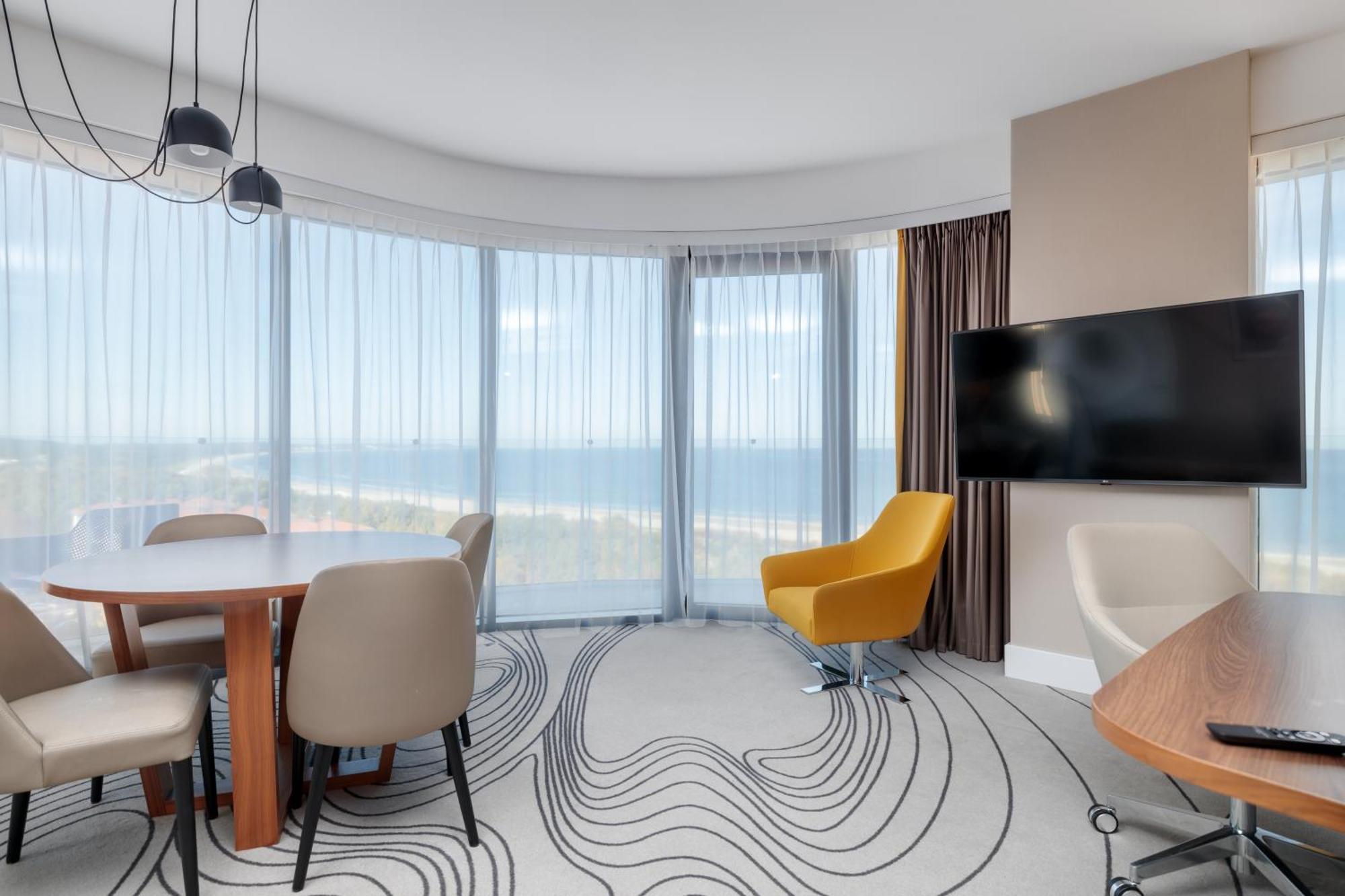 منتجع راديسون بلو سوينوجسي سفينويتشي المظهر الخارجي الصورة The image shows a modern hotel room or suite with a bright, airy atmosphere. There are large windows that allow natural light to fill the space, providing a view of the ocean. The decor includes a round dining table with several chairs, and a stylish