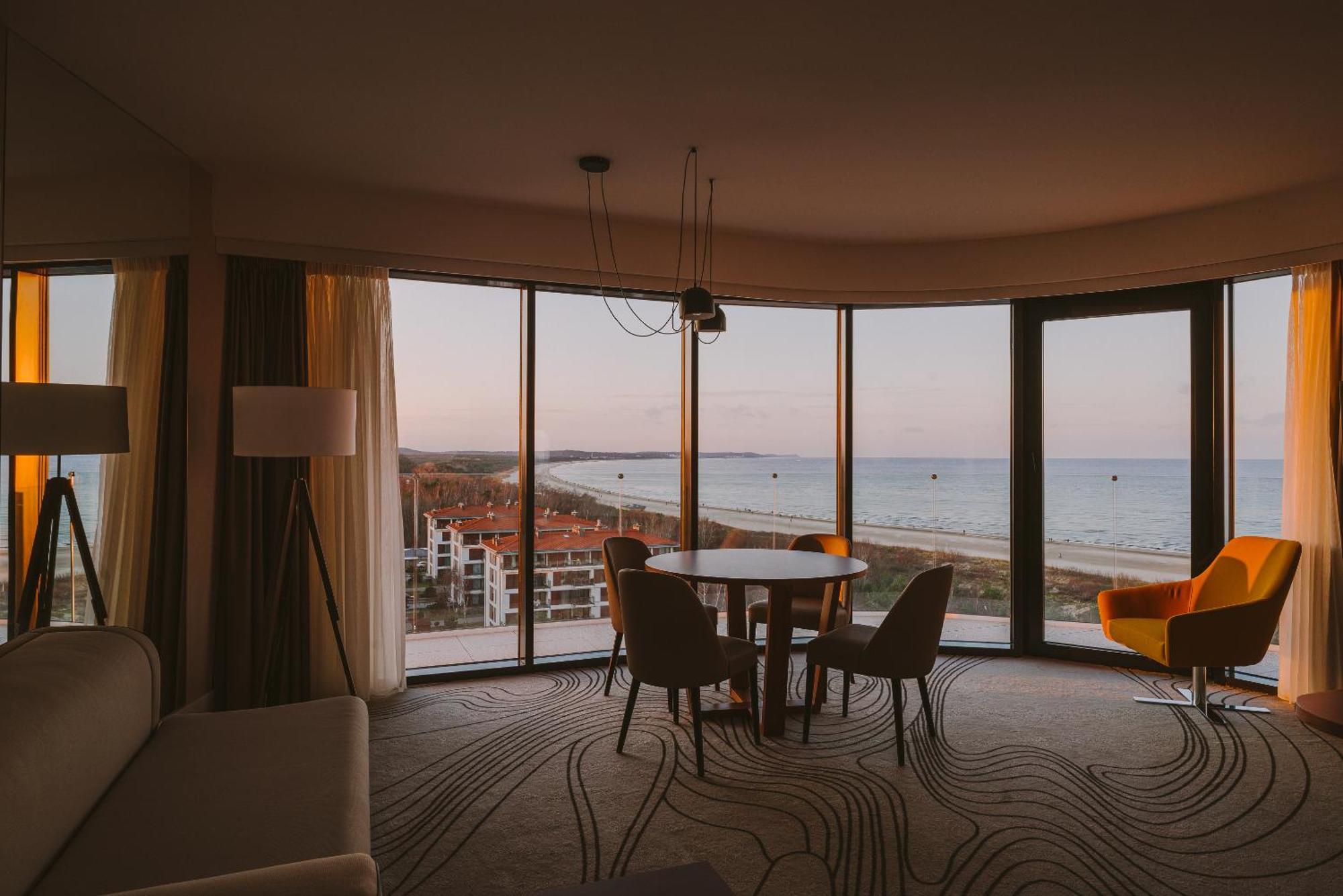 منتجع راديسون بلو سوينوجسي سفينويتشي المظهر الخارجي الصورة The photo shows a modern and stylish interior of a room featuring large windows that provide a view of the beach and ocean. The room is furnished with a round table and chairs, along with a cozy sofa and a bright yellow armchair. The decor has a cont