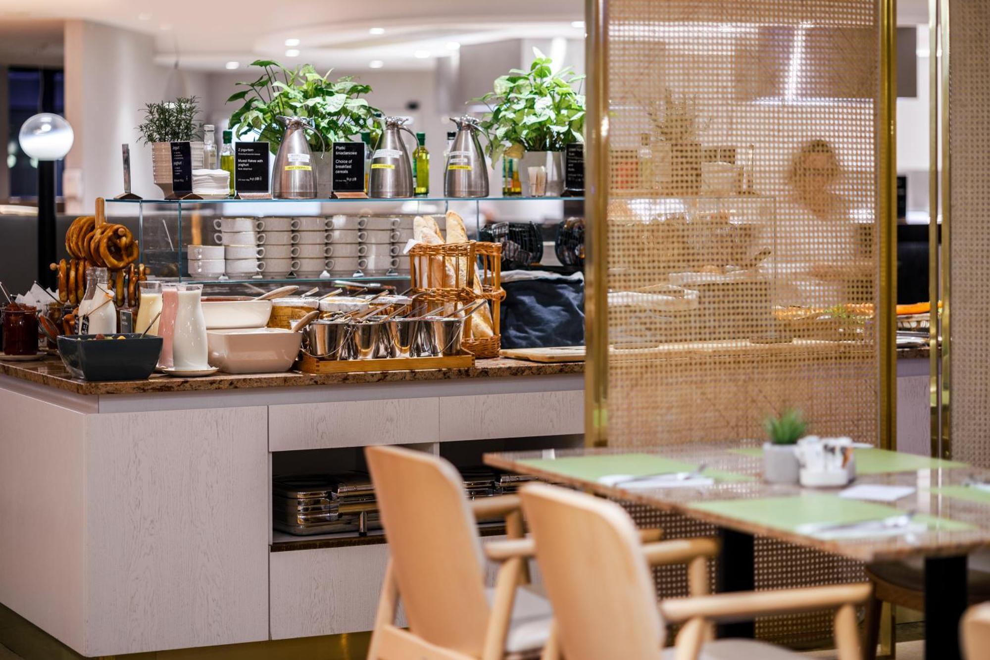 منتجع راديسون بلو سوينوجسي سفينويتشي المظهر الخارجي الصورة The photo shows a modern café or dining area. In the foreground, there are wooden chairs and tables set up for dining. In the background, there is a display counter with various items, including plants in pots, containers, and possibly dishes or uten