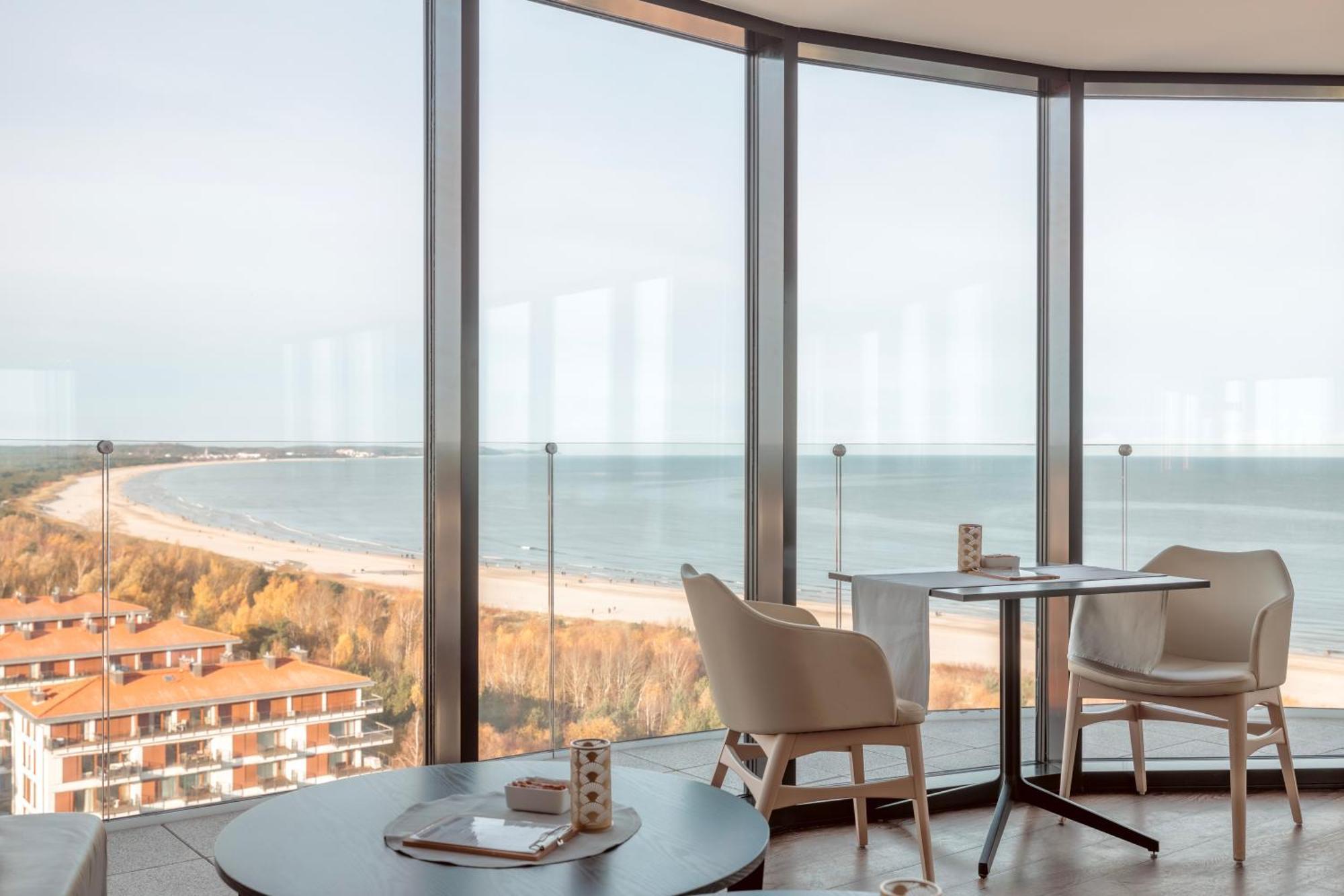 منتجع راديسون بلو سوينوجسي سفينويتشي المظهر الخارجي الصورة The photo shows a modern interior space with large floor-to-ceiling windows that provide a stunning view of the beach and sea. The area appears to be a dining or lounge space, featuring a minimalistic table and chairs. There is a sense of tranquility