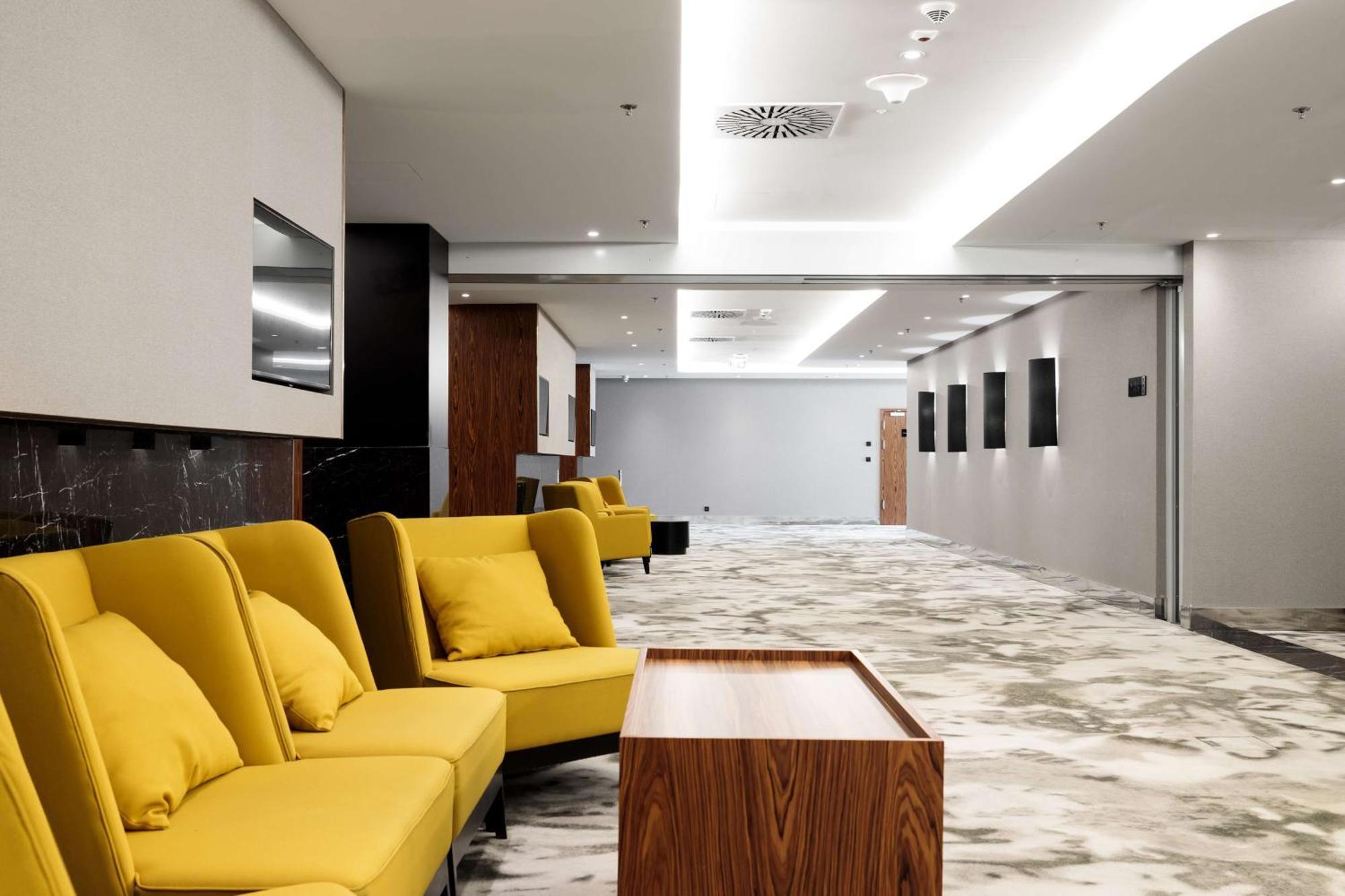 منتجع راديسون بلو سوينوجسي سفينويتشي المظهر الخارجي الصورة The photo shows a modern interior space, likely a lobby or waiting area. There are several yellow sofas with cushions arranged in a casual seating area. In front of the sofas, there is a wooden coffee table. The flooring features a gray and white mar
