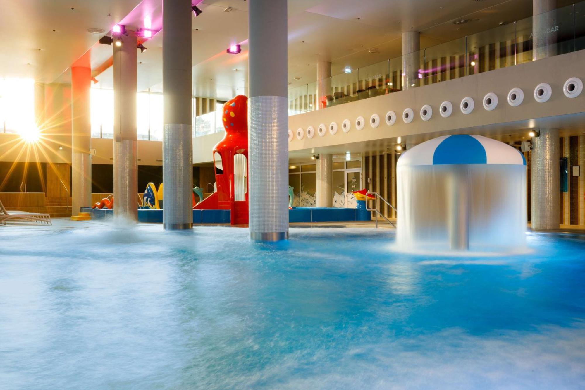 منتجع راديسون بلو سوينوجسي سفينويتشي المظهر الخارجي الصورة The photo shows an indoor water park or pool area. There is a large body of water with playful features, including a water slide shaped like a red bear and a smaller structure with a blue and white canopy. The lighting is bright, suggesting a sunny a