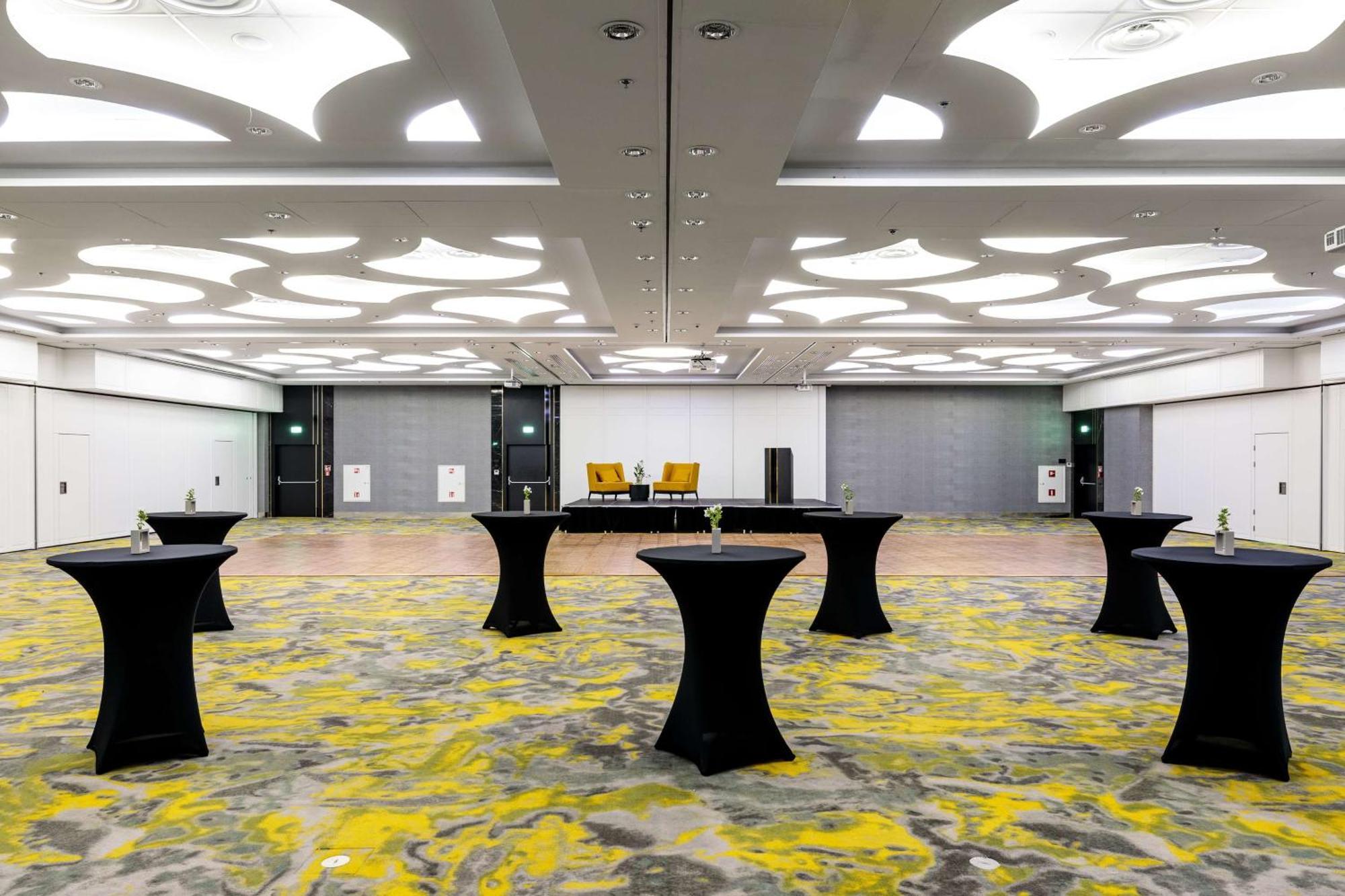 منتجع راديسون بلو سوينوجسي سفينويتشي المظهر الخارجي الصورة The photo shows a spacious interior room set up for an event. The flooring features a patterned design in gray with yellow accents. There are several black tall tables scattered throughout the area, and in the background, there are two large yellow a