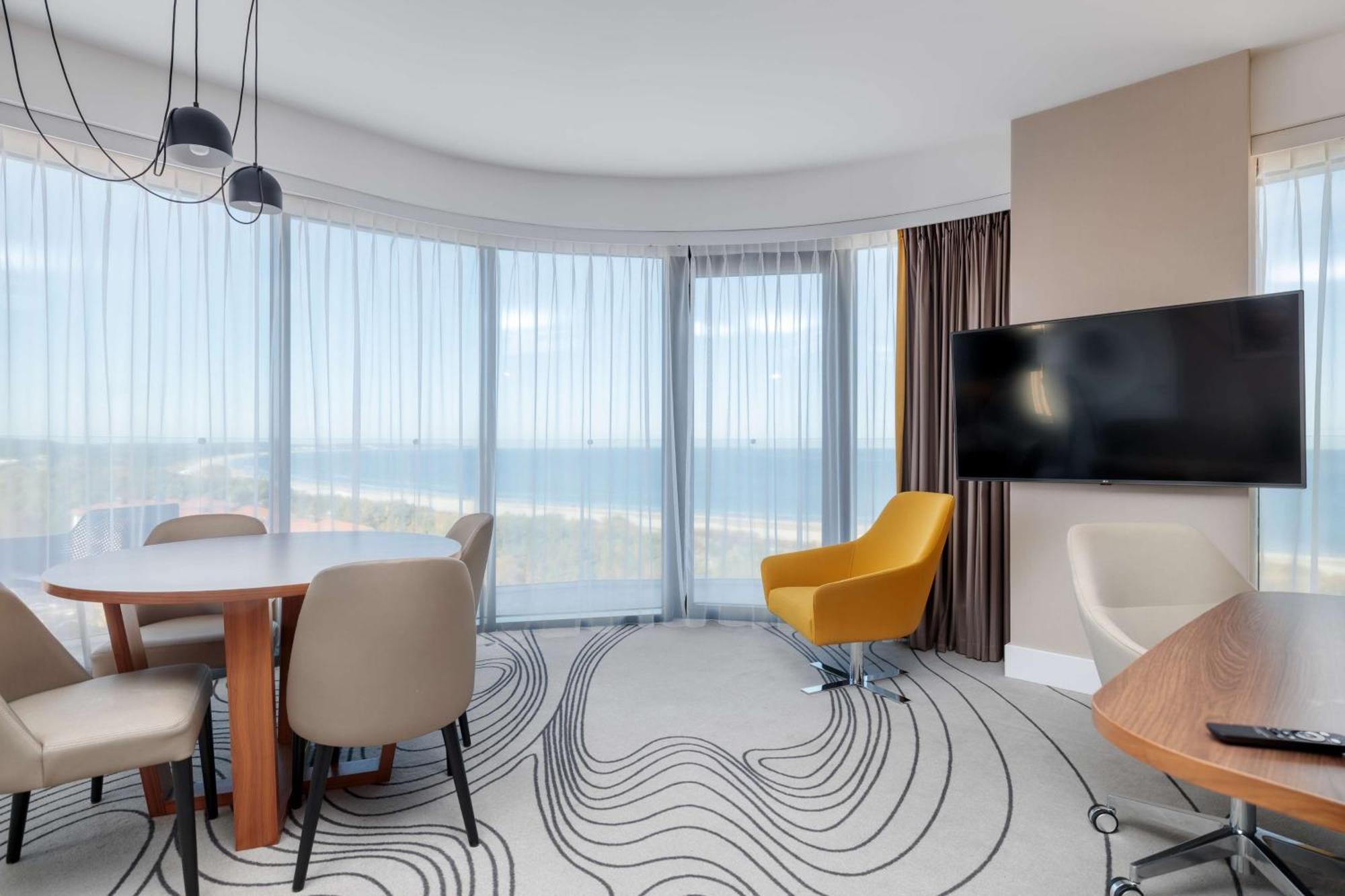 منتجع راديسون بلو سوينوجسي سفينويتشي المظهر الخارجي الصورة The photo shows a modern, stylish interior of a room with large windows that provide a panoramic view. The space features a round dining table surrounded by chairs, including a distinct yellow armchair. There is a flat-screen television mounted on th
