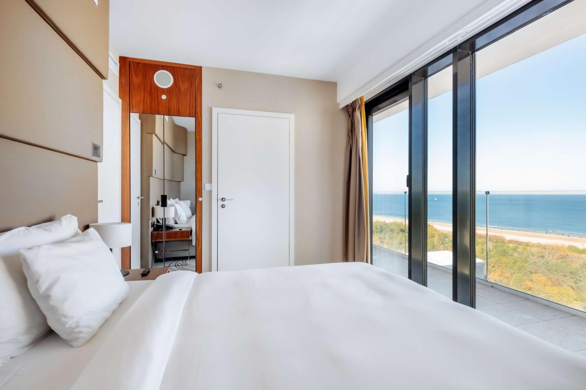 منتجع راديسون بلو سوينوجسي سفينويتشي المظهر الخارجي الصورة The photo shows a modern hotel room with a bright and airy atmosphere. There is a large bed with white bedding positioned near a glass wall that offers a view of the ocean. The room features neutral-colored walls and wooden accents, enhancing its con