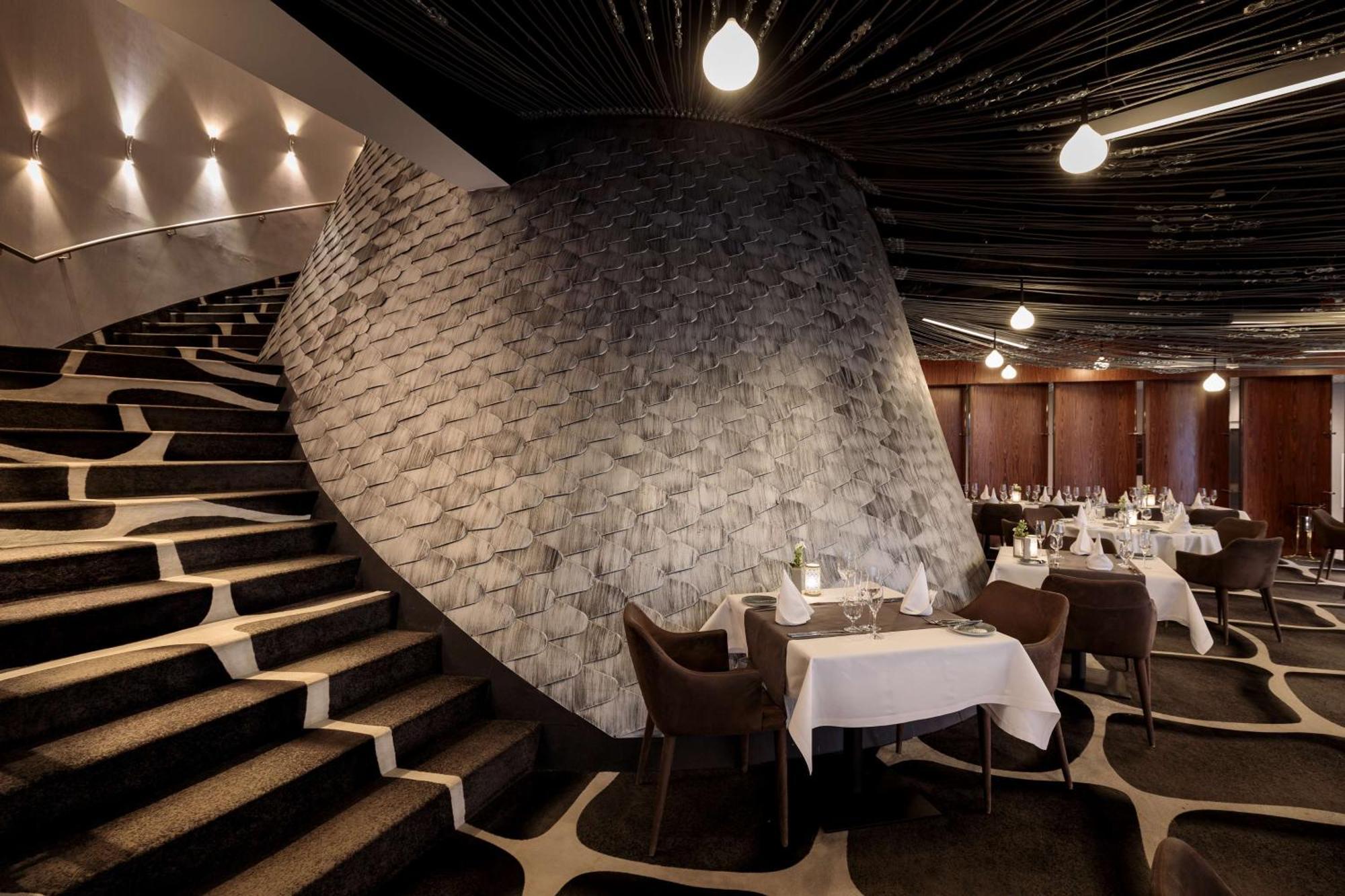 منتجع راديسون بلو سوينوجسي سفينويتشي المظهر الخارجي الصورة The photo shows the interior of a stylish restaurant. In the foreground, there are elegantly set tables with white tablecloths and dark chairs. A unique, curving stone wall adds a distinct architectural feature, and a grand staircase with a textured 