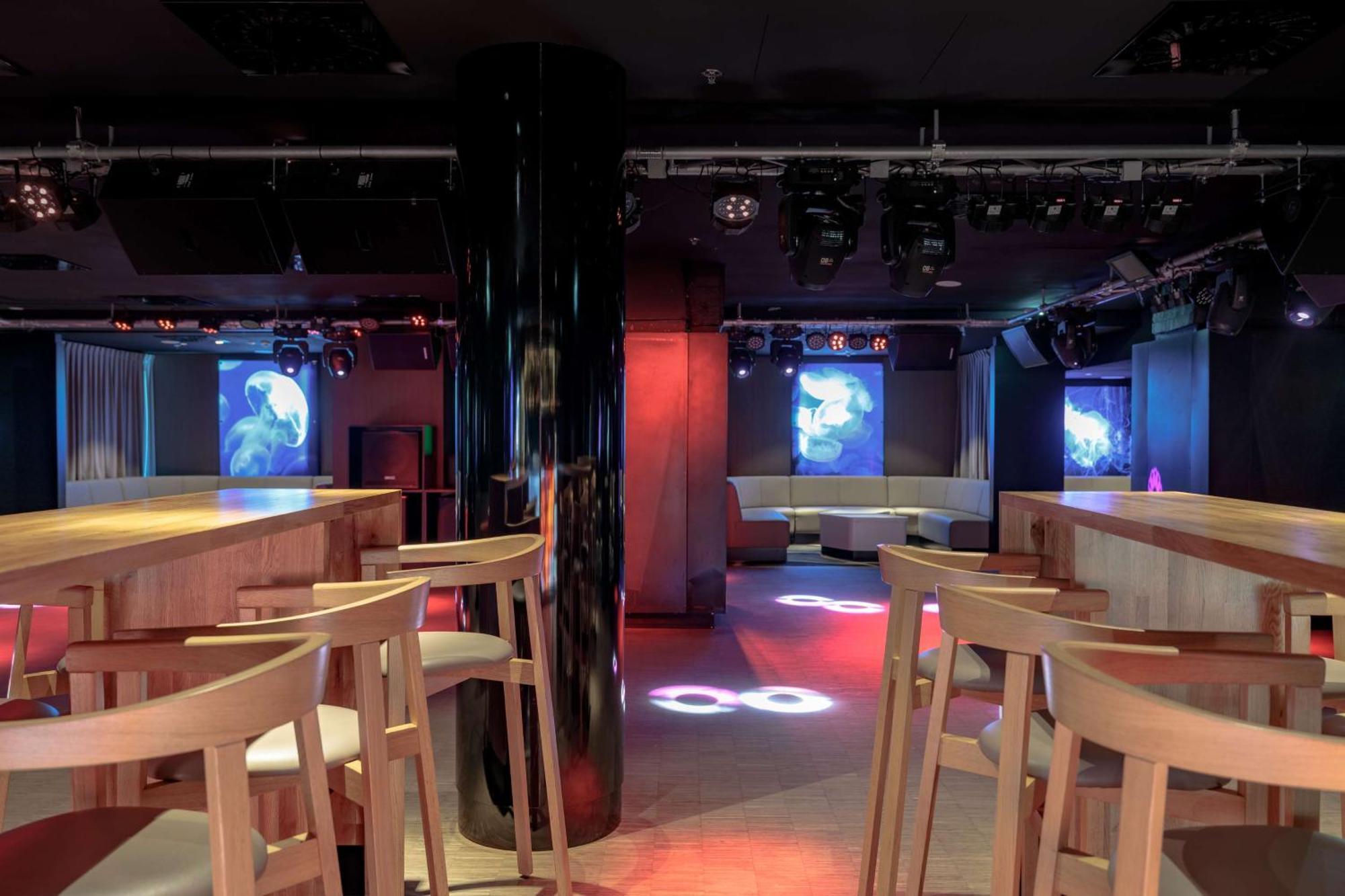 منتجع راديسون بلو سوينوجسي سفينويتشي المظهر الخارجي الصورة The photo shows an interior of a modern venue, likely a bar or club. The space features a central black column and light-colored wooden bar stools arranged around a bar area. On the walls, there are large screens displaying vibrant visuals, possibly 