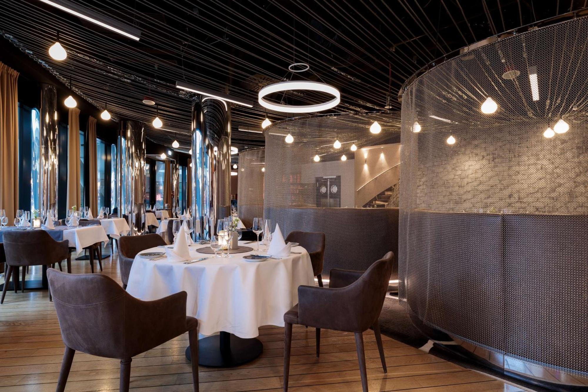 منتجع راديسون بلو سوينوجسي سفينويتشي المظهر الخارجي الصورة The image shows a modern restaurant interior that features a stylish design. There are several round tables with white tablecloths. The seating consists of brown, upholstered chairs. The space is illuminated by various hanging lights, including a dis