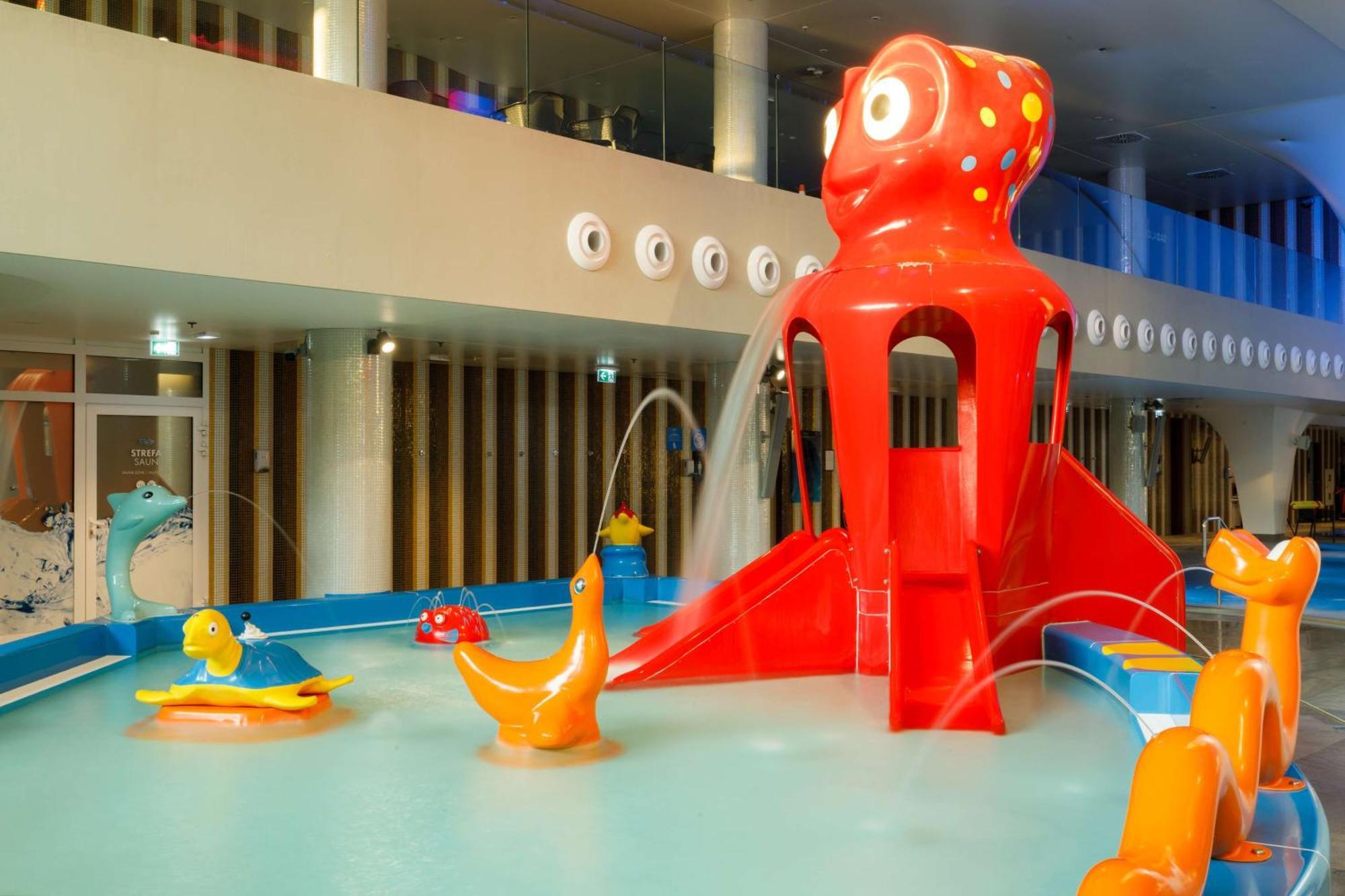 منتجع راديسون بلو سوينوجسي سفينويتشي المظهر الخارجي الصورة The photo shows a colorful indoor splash pad or water play area designed for children. Central to the scene is a large, bright red octopus structure, which has water fountains spraying out. Surrounding it are various playful, colorful water animals, 